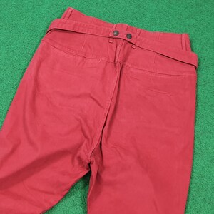 May include: Red pants with a belt detail. The pants have a high waist and a straight leg. The belt is attached to the back of the pants with two buttons. The pants are made of a solid red fabric.