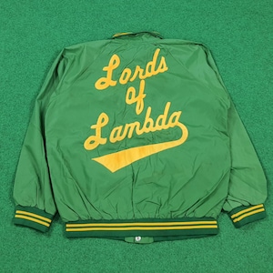 Pode incluir: Casaco bomber verde com letras amarelas que dizem "Lords of Lambda" e um design de taco de beisebol amarelo. O casaco tem punhos listrados amarelos e verdes e uma gola verde.