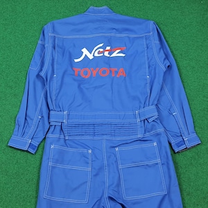 May include: A bright blue long-sleeve jumpsuit featuring white contrast stitching throughout. The back displays the white and red text "Netz" above the red text "TOYOTA". It includes a waist belt, pleated back detailing, and two large rear pockets.