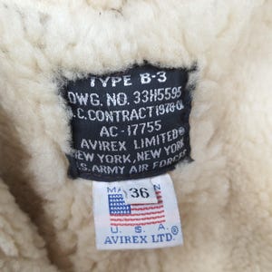 Puede incluir: Primer plano de una etiqueta negra con texto blanco y una peque&ntilde;a etiqueta con la bandera estadounidense. La etiqueta dice "TYPE B-3" y "AVIREX LIMITED&reg; NEW YORK, NEW YORK S. ARMY AIR FORCES". La etiqueta muestra la talla 36.