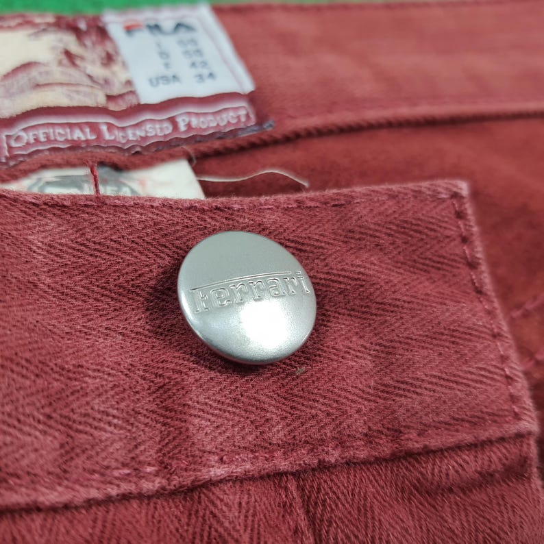 May include: Close-up of red denim pants with a silver button featuring the Ferrari logo. The pants have a textured fabric and a label that reads "USA 34" and "OFFICIAL LICENSED PRODUCT".
