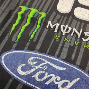 May include: A black t-shirt featuring a vertical gray striped pattern, a bright green Monster Energy claw logo, white Monster Energy text, and a large blue oval Ford logo. The graphic prints exhibit a distressed, cracked texture throughout.