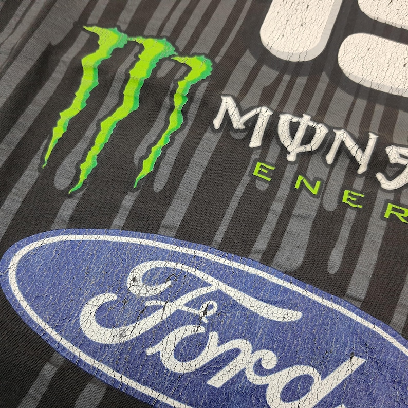 May include: A black t-shirt featuring a vertical gray striped pattern, a bright green Monster Energy claw logo, white Monster Energy text, and a large blue oval Ford logo. The graphic prints exhibit a distressed, cracked texture throughout.