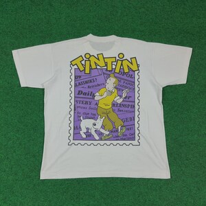 May include: White t-shirt featuring a graphic of Tintin and Snowy from the Tintin comic series. The graphic is in a purple postage stamp design with text. The shirt is on a green background.
