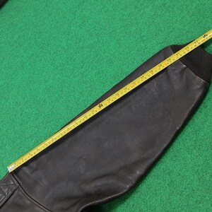 Puede incluir: Una manga de chaqueta de cuero marr&oacute;n oscuro con un pu&ntilde;o de punto acanalado negro. Una cinta m&eacute;trica amarilla est&aacute; colocada a lo largo de la manga, indicando una medida de aproximadamente 61 cm desde la costura del hombro hasta el pu&ntilde;o.