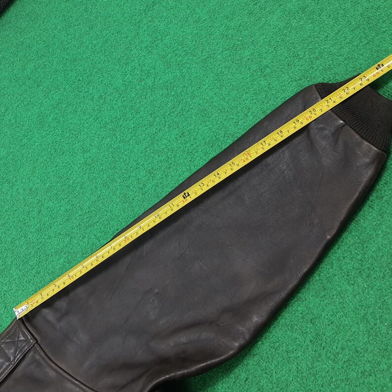Puede incluir: Una manga de chaqueta de cuero marr&oacute;n oscuro con un pu&ntilde;o de punto acanalado negro. Una cinta m&eacute;trica amarilla est&aacute; colocada a lo largo de la manga, indicando una medida de aproximadamente 61 cm desde la costura del hombro hasta el pu&ntilde;o.