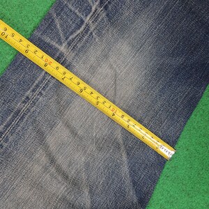 K&ouml;nnte beinhalten: Nahaufnahme von blauem Denim-Stoff mit einem gelben Ma&szlig;band. Das Band zeigt Messungen in Zentimetern, wobei die Textur und die Details des Denims hervorgehoben werden. Der Stoff liegt auf einer gr&uuml;nen Oberfl&auml;che.