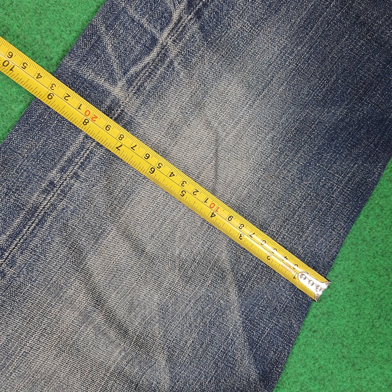 K&ouml;nnte beinhalten: Nahaufnahme von blauem Denim-Stoff mit einem gelben Ma&szlig;band. Das Band zeigt Messungen in Zentimetern, wobei die Textur und die Details des Denims hervorgehoben werden. Der Stoff liegt auf einer gr&uuml;nen Oberfl&auml;che.