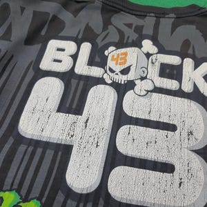 May include: A black t-shirt featuring a large, distressed white graphic print that reads BLOCK 43. The design includes a stylized skull character with the number 43 in orange on its forehead, set against a background of dark gray vertical stripes.