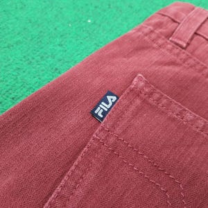May include: Close-up of burgundy-colored pants with a herringbone pattern. The back pocket and waistband are visible, with a small FILA brand label in navy blue and white.