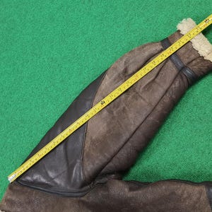 Puede incluir: Una manga de chaqueta de cuero marr&oacute;n con un panel de cuero negro, medida con una cinta m&eacute;trica amarilla. La manga tiene un forro de piel de oveja en el pu&ntilde;o. La cinta m&eacute;trica muestra una longitud de aproximadamente 61 cm.