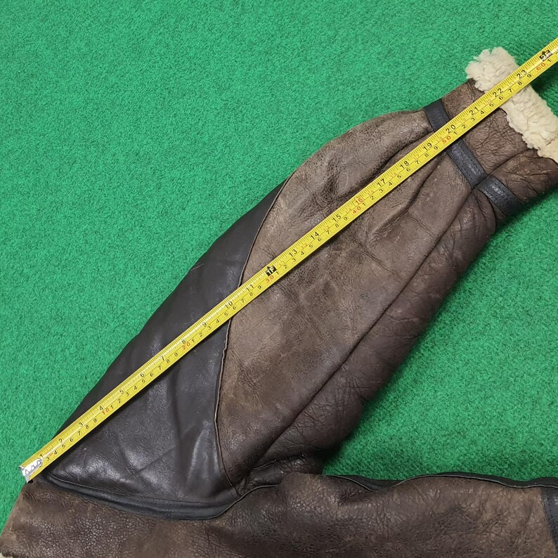 Puede incluir: Una manga de chaqueta de cuero marr&oacute;n con un panel de cuero negro, medida con una cinta m&eacute;trica amarilla. La manga tiene un forro de piel de oveja en el pu&ntilde;o. La cinta m&eacute;trica muestra una longitud de aproximadamente 61 cm.
