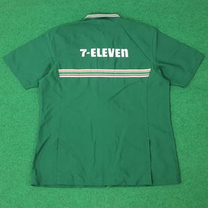 Puede incluir: Camisa de trabajo verde de manga corta con el texto "7-ELEVEN" en letras blancas en la espalda. La camisa tiene un cuello a rayas blanco y verde y una franja horizontal en rojo, blanco y naranja.
