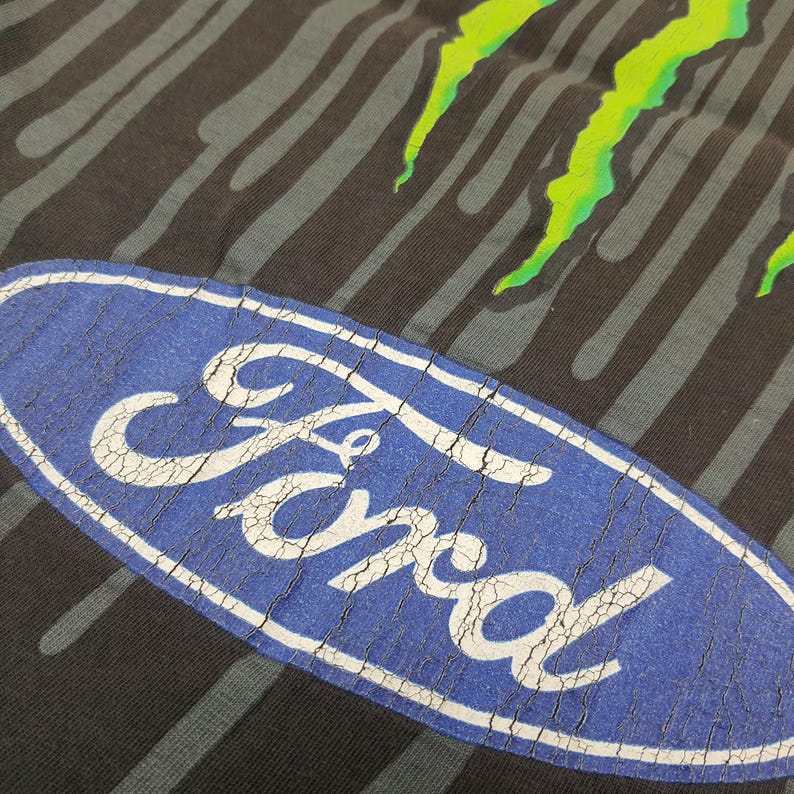 May include: A black t-shirt featuring a large blue oval Ford logo with white lettering and a cracked texture. Above the logo, a bright neon green graphic design with vertical dripping lines extends across the dark fabric background.
