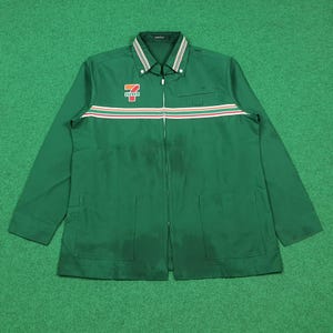 Puede incluir: Chaqueta verde con cremallera y una franja blanca, roja y naranja en el pecho. La chaqueta tiene dos bolsillos delanteros y un cuello con rayas blancas y verdes. El logotipo de 7-Eleven está en el pecho izquierdo.