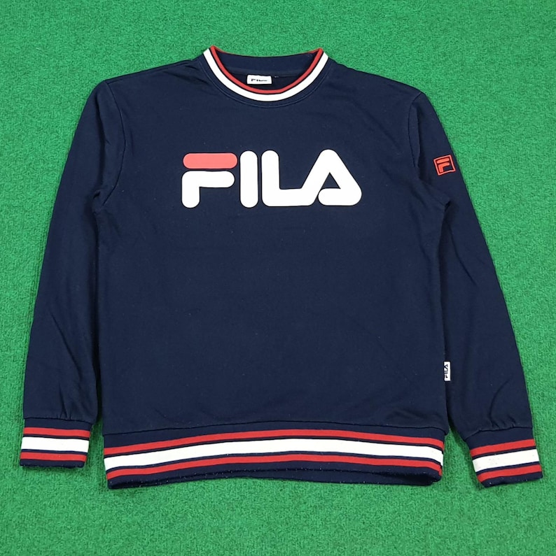 Puede incluir: Sudadera azul marino con la palabra "FILA" en letras blancas en el pecho. El logotipo de la marca tambi&eacute;n est&aacute; en la manga. El cuello, los pu&ntilde;os y el dobladillo tienen rayas rojas y blancas.