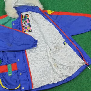 May include: A colorful vintage ski jacket with a blue base, red and green accents, and a white faux fur-lined hood. The jacket features a quilted white lining and a zipper closure. The jacket has a pocket on the inside.