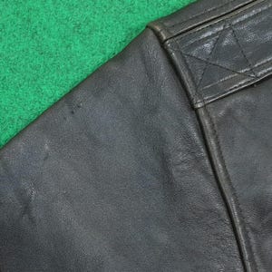 Puede incluir: Una secci&oacute;n de chaqueta de cuero marr&oacute;n oscuro con una textura suave y granulada. La prenda incluye un panel de hombro reforzado con costuras tonales visibles, mostrando un detalle de costura estructurado y un acabado de cuero mate duradero.
