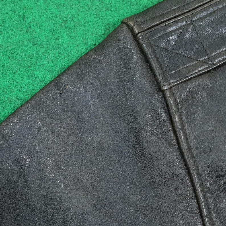 Puede incluir: Una secci&oacute;n de chaqueta de cuero marr&oacute;n oscuro con una textura suave y granulada. La prenda incluye un panel de hombro reforzado con costuras tonales visibles, mostrando un detalle de costura estructurado y un acabado de cuero mate duradero.
