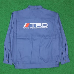 May include: A light blue long-sleeve work shirt featuring a collar, buttoned cuffs, and a large white and orange TRD Toyota Racing Development logo printed across the back. The garment has a structured design with visible vertical seams.