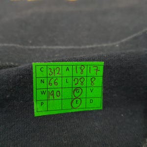May include: A black fabric surface featuring a bright green rectangular grid sticker. The sticker contains handwritten numbers and letters in black ink, including 312, 18, 17, 66, 28, 8, 190, and circled letters G and E, organized within a structured table.