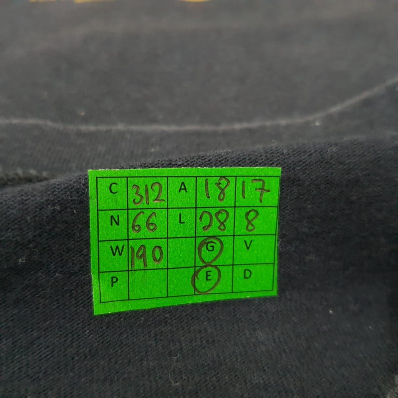 May include: A black fabric surface featuring a bright green rectangular grid sticker. The sticker contains handwritten numbers and letters in black ink, including 312, 18, 17, 66, 28, 8, 190, and circled letters G and E, organized within a structured table.