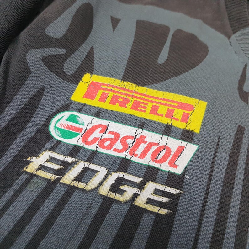 May include: A black t-shirt featuring a large, distressed graphic print with the Pirelli logo in yellow, the Castrol logo in red and green, and the word EDGE in white. The design includes vertical black stripes on a dark gray background.