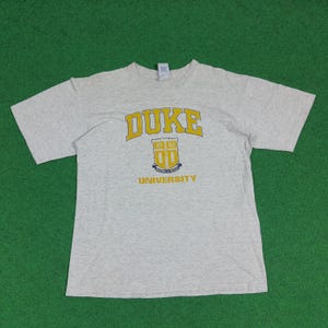 Può includere: T-shirt grigio chiaro con la scritta "DUKE" in lettere gialle sopra uno stemma universitario giallo e blu. Il testo "UNIVERSITY" è stampato sotto in giallo. La maglietta è a maniche corte.