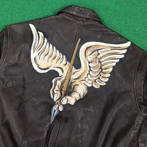 Puede incluir: Una chaqueta de cuero marr&oacute;n oscuro con un gran dise&ntilde;o pintado a mano en la espalda. La ilustraci&oacute;n muestra una mano alada estilizada que sostiene una pluma, representada en tonos crema, beige y gris, centrada en el panel posterior de la prenda.