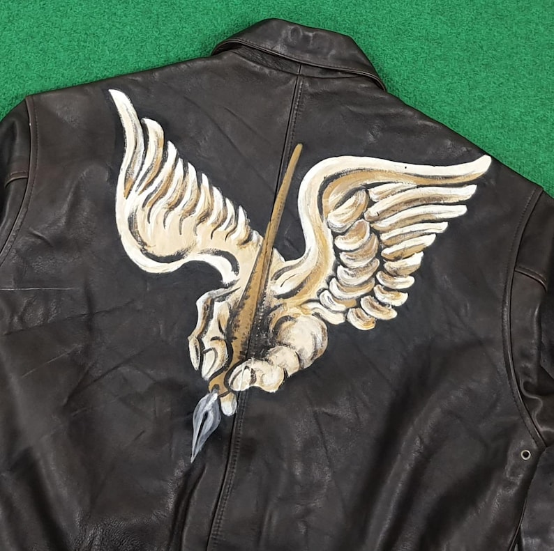 Puede incluir: Una chaqueta de cuero marr&oacute;n oscuro con un gran dise&ntilde;o pintado a mano en la espalda. La ilustraci&oacute;n muestra una mano alada estilizada que sostiene una pluma, representada en tonos crema, beige y gris, centrada en el panel posterior de la prenda.