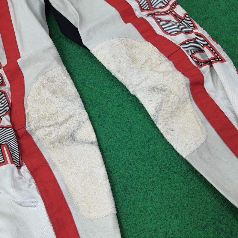 May include: Close-up of a white and red racing suit with padded knee protection. The suit has red stripes and black and white graphic details. The knee pads are off-white and show signs of wear. The background is green.