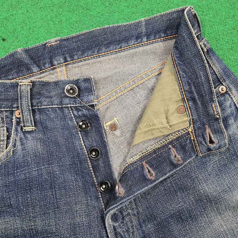 K&ouml;nnte beinhalten: Nahaufnahme einer blauen Jeans mit Knopfleiste. Die Jeans hat schwarze Kn&ouml;pfe und sichtbare N&auml;hte in einem kontrastierenden gelben Faden. Der Hosenschlitz ist teilweise aufgekn&ouml;pft und gibt den Blick auf den Innenstoff frei.