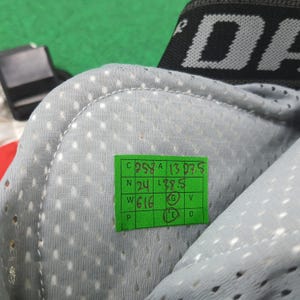 May include: Close-up of a gray, perforated fabric with a stitched edge. A green sticker displays numbers and letters: C 258 A 13 07 5, N 24, L 38.5, W 616, and other letters. A black strap with the letters "DA" is visible.