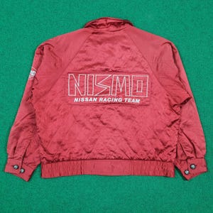May include: A red satin jacket with the text "NISMO NISSAN RACING TEAM" in white. The jacket has a quilted pattern, a collar, and buttoned cuffs. The jacket is a vintage racing style.