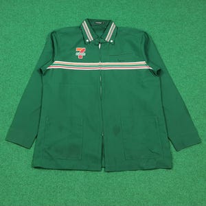 Puede incluir: Chaqueta de trabajo verde con cremallera completa, dos bolsillos delanteros y cuello. La chaqueta presenta un diseño de rayas horizontales en rojo, blanco y marrón en el pecho. El logotipo de "7-Eleven" está en el pecho izquierdo.