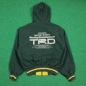May include: A dark green hooded jacket featuring the white text Toyota Motor Sports Racing Development TRD on the back. The jacket has long sleeves with yellow and green striped ribbed cuffs and a matching ribbed hem, plus a yellow inner lining.