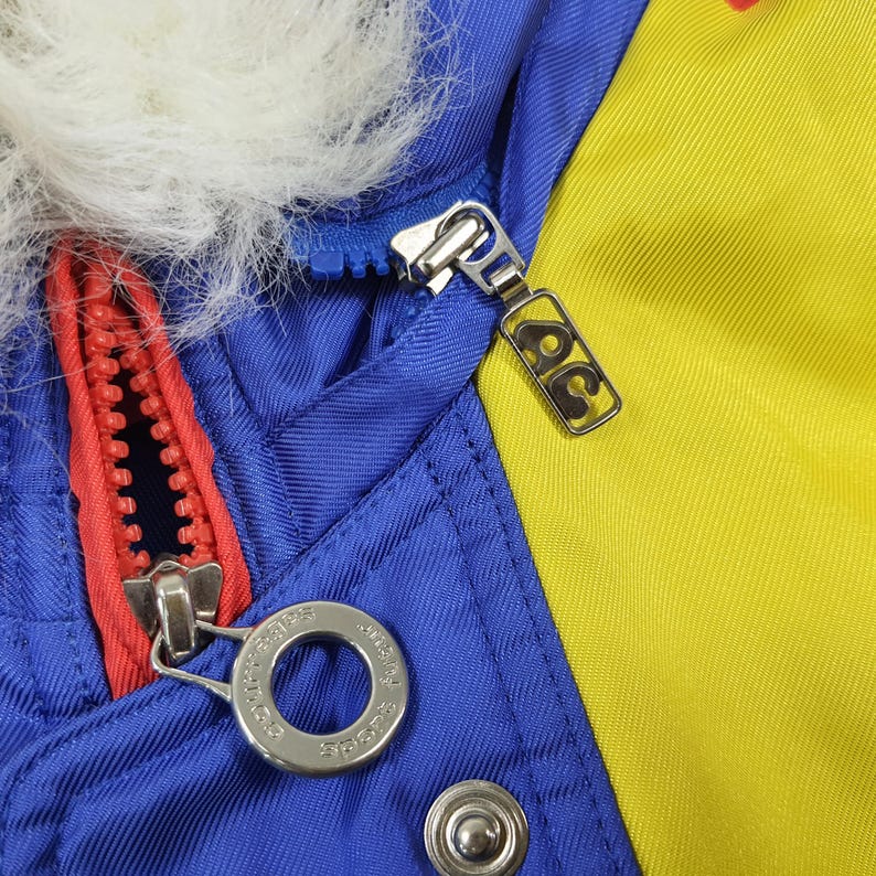 May include: Close-up of a blue, yellow, and red jacket with a white fur-lined hood. The jacket features a red zipper and a silver zipper pull. The jacket has a silver button and a silver zipper pull with the letters "AC" on it.