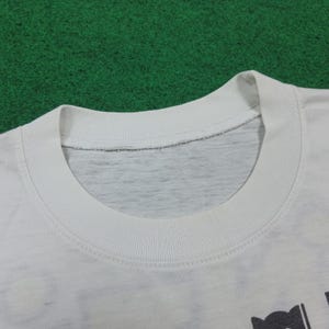 May include: Close-up of a white t-shirt collar. The collar is ribbed and the neckline is a lighter gray color. The shirt appears to be made of a soft, comfortable material. The shirt is on a green surface.