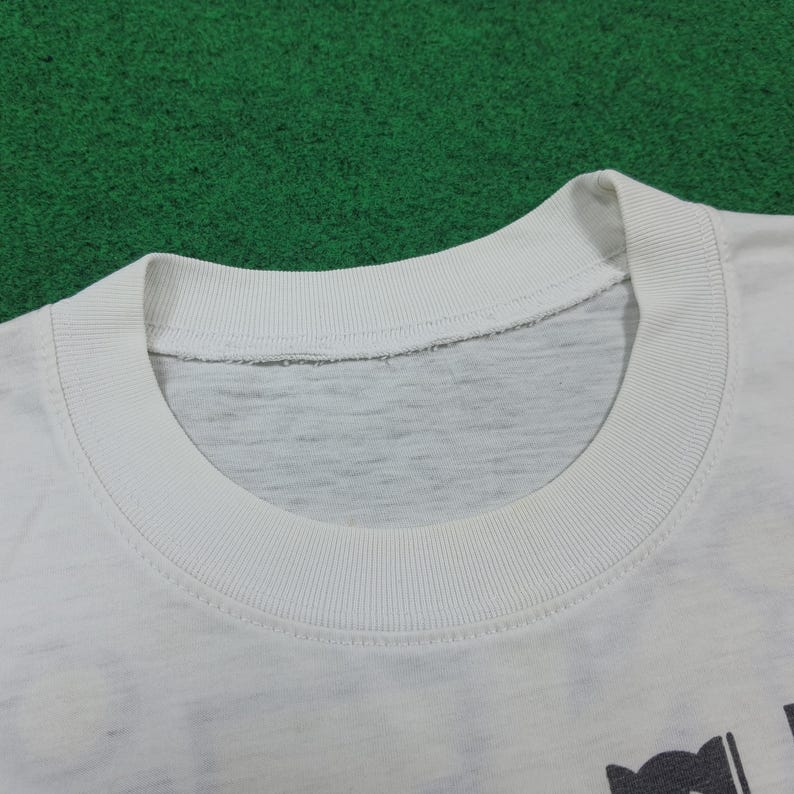 May include: Close-up of a white t-shirt collar. The collar is ribbed and the neckline is a lighter gray color. The shirt appears to be made of a soft, comfortable material. The shirt is on a green surface.