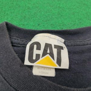 May include: A dark navy blue t-shirt featuring a white fabric neck label with the black and yellow CAT logo printed on it. The label includes the text MADE IN U.S.A. and MEDIUM, indicating the size of the garment and its country of origin.