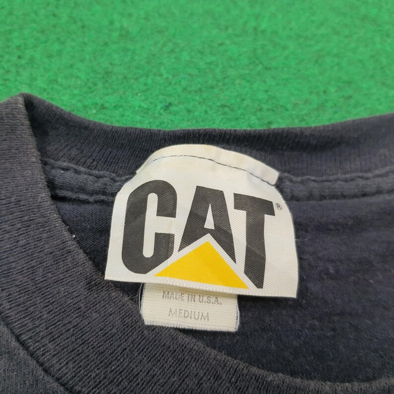 May include: A dark navy blue t-shirt featuring a white fabric neck label with the black and yellow CAT logo printed on it. The label includes the text MADE IN U.S.A. and MEDIUM, indicating the size of the garment and its country of origin.
