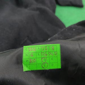 K&ouml;nnte beinhalten: Nahaufnahme eines schwarzen Kleidungsst&uuml;cks mit einem angebrachten gr&uuml;nen Etikett. Das Etikett enth&auml;lt handschriftlichen Text und Zahlen, darunter "C 5526 A 2318" und "L 26 04.5". Der Stoff des Kleidungsst&uuml;cks scheint glatt und leicht reflektierend.