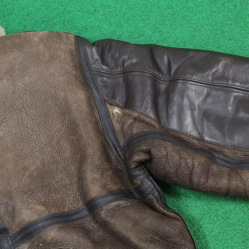 Puede incluir: Primer plano de una manga de chaqueta de cuero marr&oacute;n con la parte superior del brazo de cuero marr&oacute;n oscuro. La chaqueta tiene ribetes negros y una superficie texturizada. El fondo es verde.