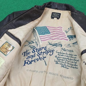 Puede incluir: Una chaqueta de vuelo de cuero marr&oacute;n oscuro con un forro interior de color crema. El forro presenta una bandera estadounidense impresa, ilustraciones de aviones antiguos y el texto: The Stars and Stripes Forever, Mother y Avirex Ltd.