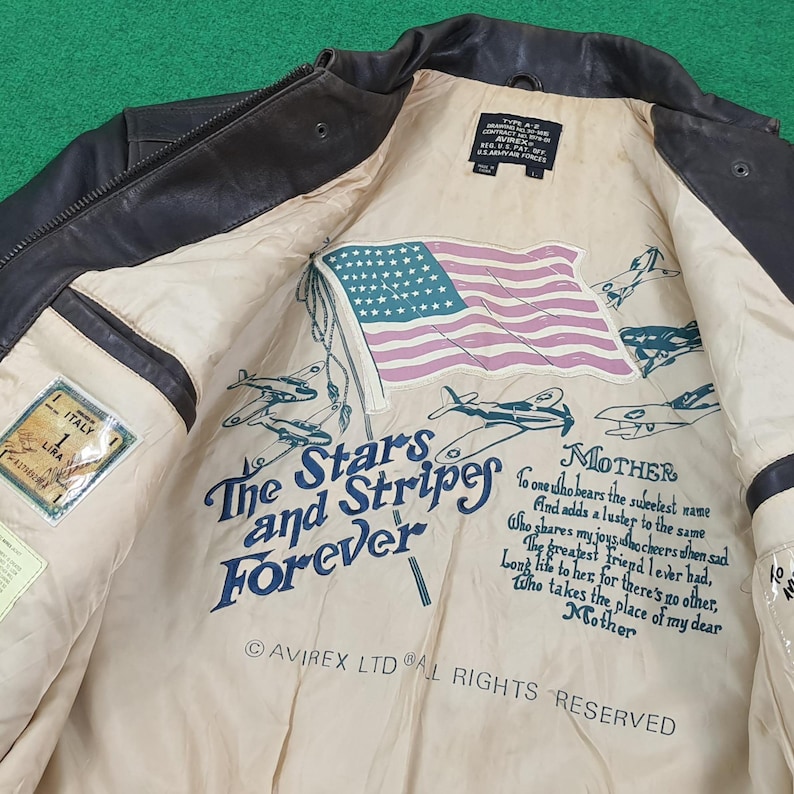 Puede incluir: Una chaqueta de vuelo de cuero marr&oacute;n oscuro con un forro interior de color crema. El forro presenta una bandera estadounidense impresa, ilustraciones de aviones antiguos y el texto: The Stars and Stripes Forever, Mother y Avirex Ltd.