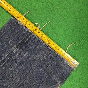 K&ouml;nnte beinhalten: Nahaufnahme eines Abschnitts einer blauen Jeans mit ausgefranstem Rand. Ein gelbes Ma&szlig;band liegt auf dem Stoff und zeigt Messungen in Zentimetern an. Die Jeans befindet sich auf einer gr&uuml;nen Oberfl&auml;che.