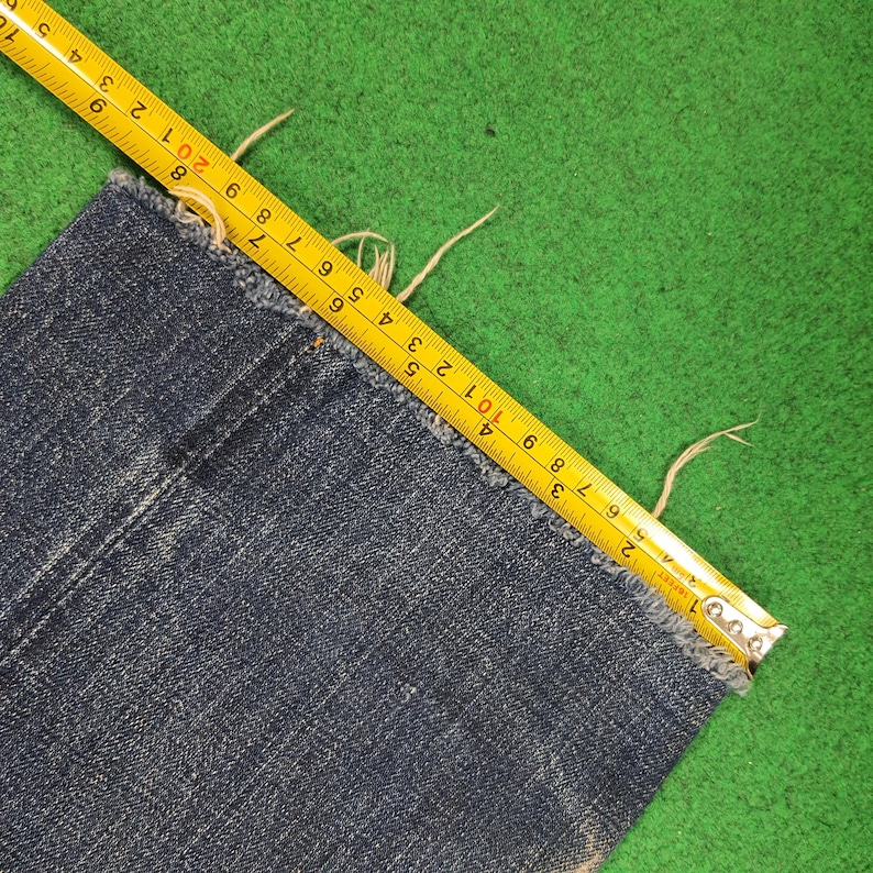 K&ouml;nnte beinhalten: Nahaufnahme eines Abschnitts einer blauen Jeans mit ausgefranstem Rand. Ein gelbes Ma&szlig;band liegt auf dem Stoff und zeigt Messungen in Zentimetern an. Die Jeans befindet sich auf einer gr&uuml;nen Oberfl&auml;che.
