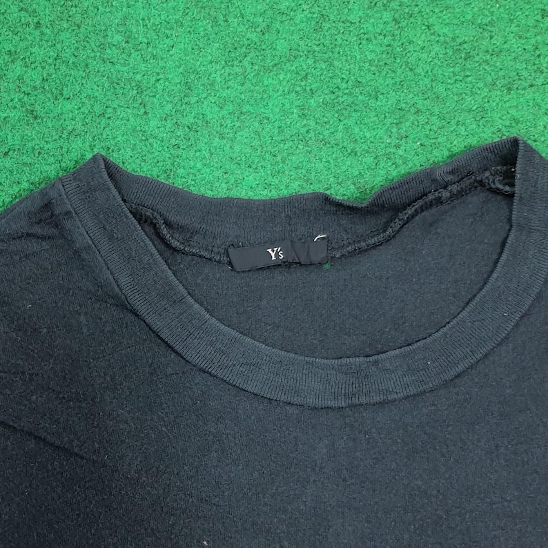 May include: A black crew neck t-shirt featuring a ribbed collar and a small black fabric label sewn into the inner neckline. The label displays the white text Y's. The garment is made of a soft, dark fabric with a visible texture throughout the material.