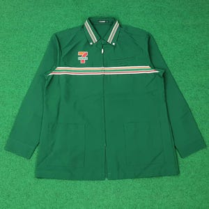 Puede incluir: Chaqueta verde con cremallera, mangas largas y dos bolsillos delanteros. La chaqueta presenta una franja blanca, roja y naranja en el pecho y el logotipo "7-ELEVEN" en el pecho izquierdo. El cuello tiene un diseño a rayas blancas y verdes.