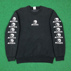 Może przedstawiać: Czarna bluza z okrągłym dekoltem z białą grafiką czaszki nad napisem SKATES wyśrodkowanym na klatce piersiowej. Każdy długi rękaw posiada pionową kolumnę pięciu identycznych białych grafik czaszek z napisem SKATES umieszczonym poniżej.